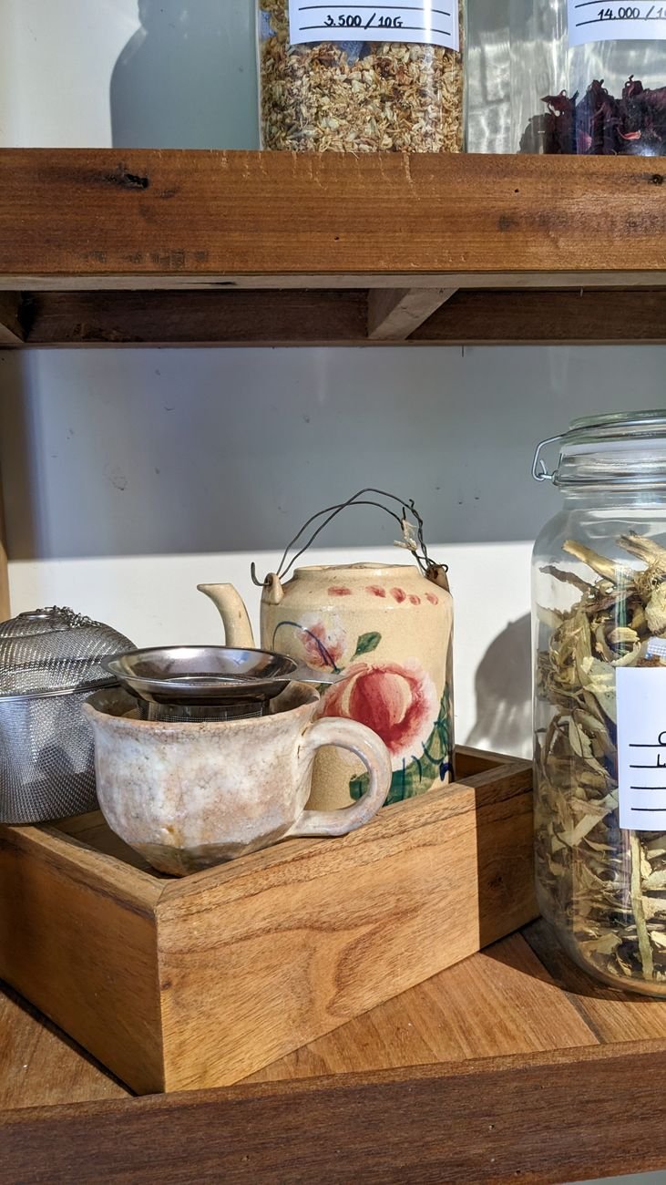 Rustic arrangement featuring a floral ceramic teapot, cup, and jars of herbs — botanical morning ritual ingredients